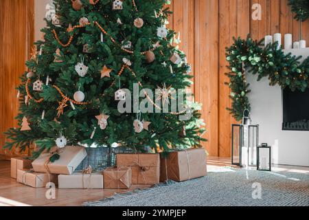 Wunderschön geschmückte Weihnachtsbaumstände in der Nähe des Kamins, dekoriert mit Girlande auf warmem hölzernem Hintergrund, umgeben von eingewickelten Geschenkboxen. Fest Stockfoto