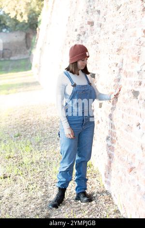 Modische junge Frau, die Overalls und Mütze trägt und eine alte Ziegelmauer in einem Park berührt Stockfoto