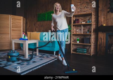 Foto von positiver guter Laune Frau gekleidetes weißes Hemd, die Hausarbeit macht und Selfie macht, modernes Gadget im Wohnraum des Hauses Stockfoto