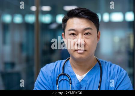 Asiatischer Mann Arzt in blauen Peelings steht selbstbewusst mit Stethoskop in moderner Gesundheitseinrichtung. Ausdruck ernst, Projizierung von Professionalität und Hingabe im medizinischen Bereich. Stockfoto
