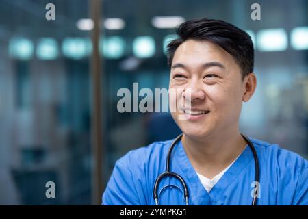 Asiatischer Mann Arzt in blauer Uniform lächelt selbstbewusst im Krankenhauskorridor. Das Bild zeigt Wärme, Professionalität, Engagement für die Gesundheitsversorgung. Stockfoto