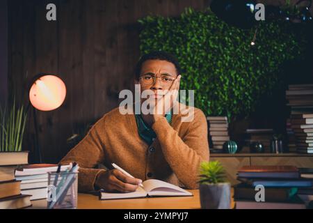 Junge Studentin im lässigen Pullover, die am späten Abend am Schreibtisch studiert, umgeben von Büchern und Pflanzen unter natürlichem Licht. Stockfoto