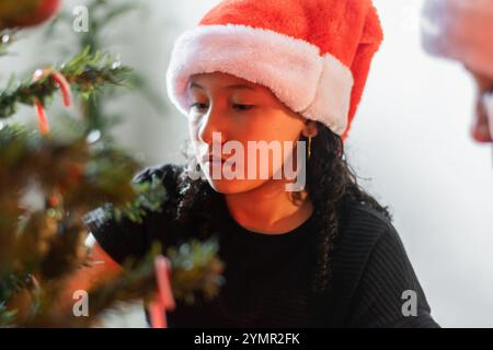 Nahaufnahme eines kleinen lateinischen Mädchens mit einem nachdenklichen und konzentrierten Gesicht neben dem weihnachtsbaum, mit einem weihnachtsmütze. Stockfoto