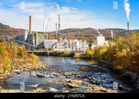 Papiermühle mit hohen Schornsteinen, die im Herbst weißen Rauch am Ufer eines Flusses in einer bewaldeten Berglandschaft ausstoßen Stockfoto