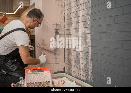 Ein Mann, der Arbeitskleidung trägt und eine Malerwalze hält, wird beobachtet, wie er sorgfältig Farbe auf eine Ziegelwand aufträgt. Der Mann konzentriert sich auf die Stockfoto