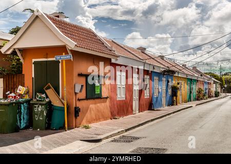 Eine Reihe kleiner, einstöckiger Häuser aus der Kolonialzeit in Terrakotta, Rot, Blau und Grün am Fort Nassauweg in Willemstad, Curacao. Diese einfachen Arbeiterhäuser stehen im Kontrast zu den raffinierteren historizistischen Fassaden, die in anderen Teilen der Stadt zu finden sind. Die Lichter deuten darauf hin, dass die Straße für Festivals oder Märkte genutzt wird. Farbenfrohe Fassaden an renovierten Häusern in der Bargestraat zwischen Schrloo und Fleur de Marie, die mit hellen Dekorationen bedeckt ist, Kòrsou Stockfoto