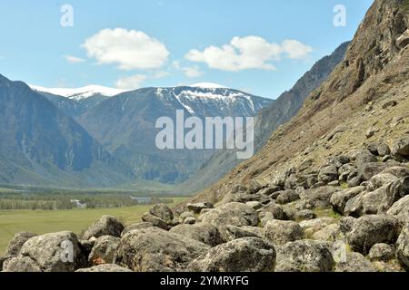 Eine Verstreuung großer Steine am Fuße einer Klippe mit Blick auf ein malerisches Tal, umgeben von schneebedeckten Bergketten. Chulyshman River vall Stockfoto