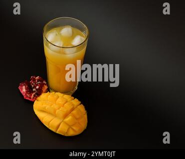Granatapfel-Mangobaumsaft in einem hohen facettierten Glas mit Eis auf schwarzem Hintergrund, neben reifem Obst. Nahaufnahme. Stockfoto
