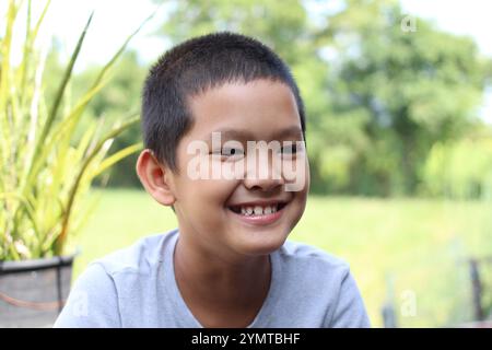 Ein fröhlicher kleiner Junge lächelt hell in einer natürlichen Umgebung im Freien, umgeben von üppigem Grün. Sein Ausdruck spiegelt Glück und Unschuld wider. Stockfoto