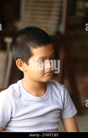 Ein kleiner Junge mit nachdenklichem Ausdruck sitzt drinnen und trägt ein graues T-Shirt. Sein ruhiges Verhalten und seine entspannte Haltung vermitteln einen Moment ruhiger Introspe Stockfoto