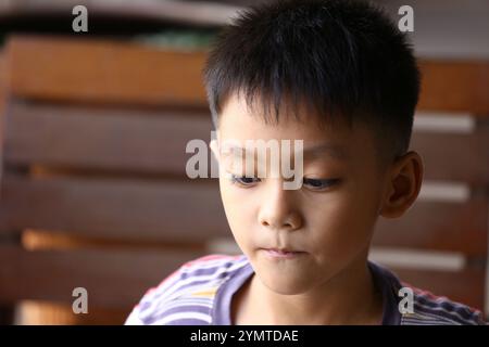 Ein nachdenklicher kleiner Junge mit kurzen Haaren sitzt drinnen und blickt mit einem ernsten Ausdruck nach unten. Sein gestreiftes Hemd verleiht einem ruhigen Moment einen Hauch von Farbe. Stockfoto