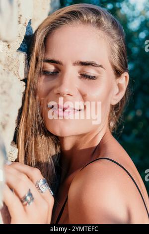 Frau, Porträt, Lächeln - Nahaufnahme Porträt einer lächelnden Frau mit langen blonden Haaren, die an eine Steinmauer gelehnt sind. Stockfoto