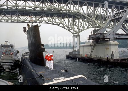 Besatzungsmitglieder stehen an Bord des nuklearbetriebenen Angriffs-U-Bootes Ex-USS Nautilus (SSN 571), das 1994 unter einer Brücke zur U-Boot-Basis New London, Conecticut USA, gezogen wird. Der große Hafenschlepper Negwagon (YTB 834) hilft am Hafenbogen des U-Bootes. Stockfoto