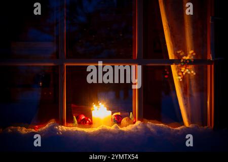 Nachtfenster eines Holzhauses in einem Winterwald. Weihnachtskerzen und -Dekorationen sind durch das Fenster sichtbar Stockfoto