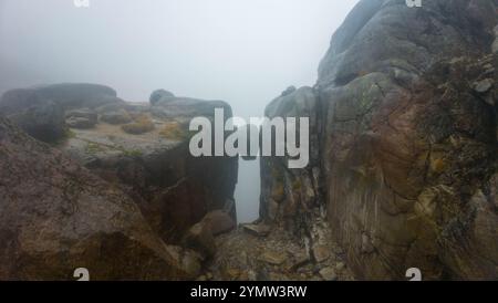 Eine nebelige Landschaft mit einem großen Felsbrocken zwischen zwei Klippen, umgeben von rauem Gelände und Nebel. Stockfoto