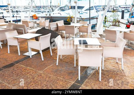 Sitzbereich im Freien an einem Yachthafen mit Tischen und Stühlen auf gefliestem Boden. Im Hintergrund legen Boote an der Marina an und fügen eine hinzu Stockfoto