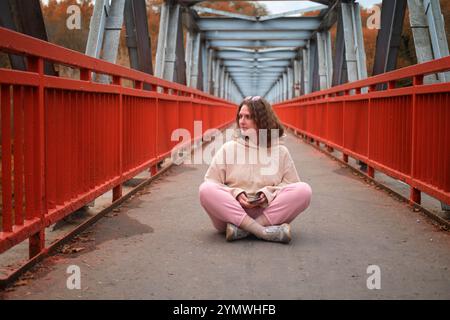 Ein einsamer Mensch sitzt friedlich auf einer riesigen Brücke und umhüllt die kalte Herbstluft. Die erwachsene Frau in Herbstkleidung sitzt auf einer Industriebrücke Stockfoto