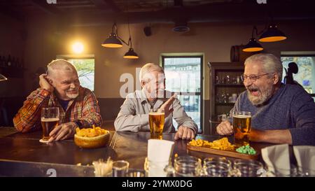 Positive ältere Frauen treffen sich in einer warm beleuchteten Bar, trinken Bier mit Snacks, reden und lachen. Freundliche kleine Unterhaltung und Freizeit Stockfoto