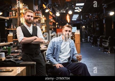 Brutaler, selbstbewusster Mann, der mit seinem neuen Look zufrieden ist. Erfahrener Friseur, der das Ergebnis seiner Arbeit zeigt. Zwei bärtige Männer lächeln vor die Kamera. Stockfoto