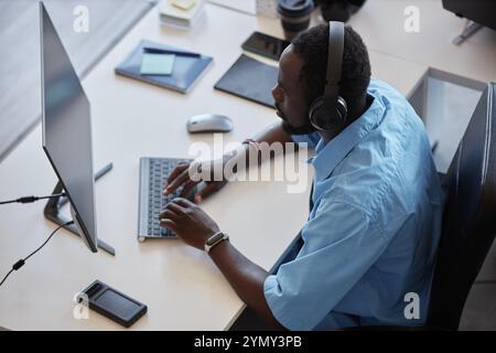 Afroamerikanischer Mann mit Kopfhörern, arbeitet am Computer an einem modernen Bürotisch mit diversen Zubehörteilen, zeigt eine Ansicht von oben nach unten mit Profi Stockfoto