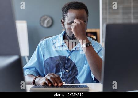 Mann in einem blauen Hemd, der am Schreibtisch sitzt und den Kopf in Gedanken kratzt, während die Zimmereinrichtung im Hintergrund sichtbar ist. Fokussierter Ausdruck auf fac Stockfoto