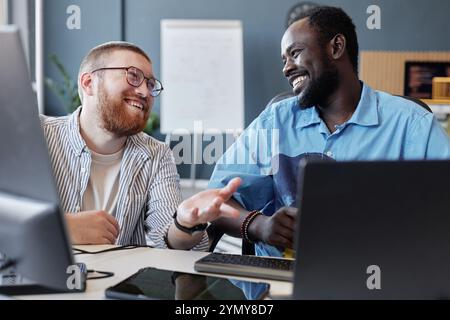 Zwei Kollegen sitzen an einem Schreibtisch, während sie lächeln und über die Arbeit diskutieren. Laptops, Dokumente und andere Büromaterialien sind in modernen Arbeitsbereichen sichtbar Stockfoto