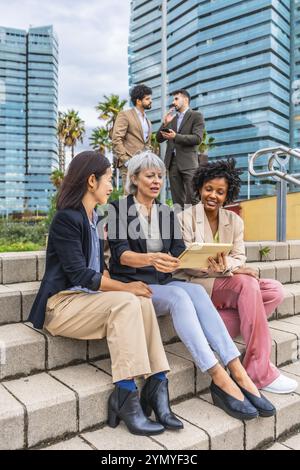 Vertikales Foto von drei multiethnischen Geschäftsfrauen, die auf einer Freitreppe sitzen und mit einem digitalen Tablet arbeiten Stockfoto