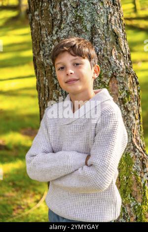 Vertikales Porträt eines kaukasischen Jungen, der mit überkreuzten Armen in der Natur neben Baumstamm in einem Alpenwald steht Stockfoto