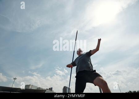 Athlet übt Stangensprung auf dem Feld im Freien mit Sichtbarkeit der Stadt im Hintergrund. Die Action wird während des sonnigen Tages aufgenommen, während der Athlet in der Luft nicht in die Kamera schaut Stockfoto