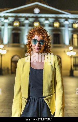 Frau mit lockigen Haaren in eleganter Freizeitkleidung, die nachts in der Stadt steht Stockfoto