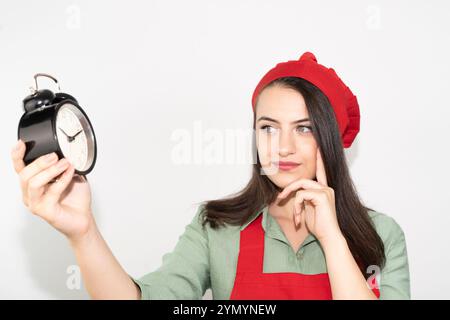 Junge nachdenkliche Hausfrau in Schürze, kochende Bäckerin, die Uhr hält und denkt Stockfoto