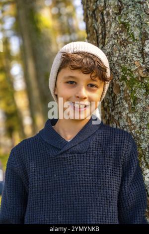 Vertikales Nahporträt eines städtischen Teenagers mit Hut in einem Baumstamm, der im Wald in die Kamera lächelt Stockfoto