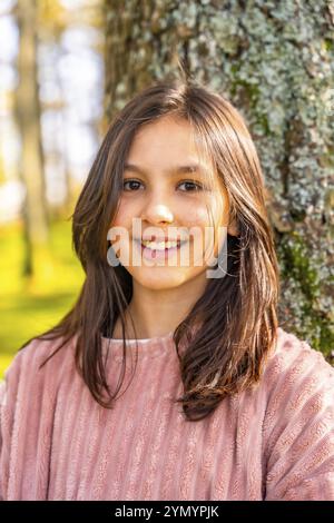 Nahaufnahme eines vertikalen Porträts eines kaukasischen lächelnden Mädchens mit rosa Kleidung, das die Natur genießt und sich auf einen Baum stützt Stockfoto