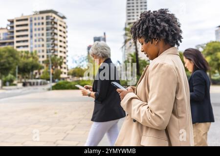 Seitenansicht multiethnischer Geschäftsleute, die in ihre Mobiltelefone vertieft sind und durch die Stadt laufen Stockfoto