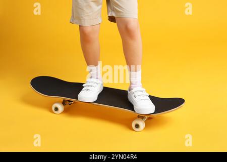 Kleines Mädchen, das auf dem Skateboard steht, vor gelbem Hintergrund, Nahaufnahme Stockfoto