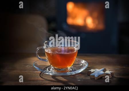 Dampfender heißer Tee in einer Glastasse und Zimt-Sternenkekse auf einem rustikalen Holztisch vor einem Kamin, gesundes Getränk in einem gemütlichen Zuhause im Winter oder Stockfoto