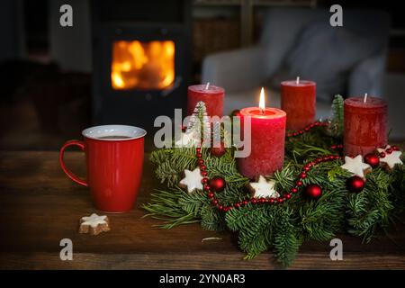 Adventskranz mit Weihnachtsdekoration und roten Kerzen, die erste angezündet, und eine Tasse Kaffee auf einem rustikalen Tisch vor dem Kamin in einer gemütlichen Stockfoto