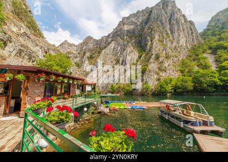 Touristen, die eine malerische Bootsfahrt auf dem smaragdgrünen Wasser des Matka Canyon genießen, mit Restaurants und der Kirche des Klosters St. Andrea in Mazedonien Stockfoto
