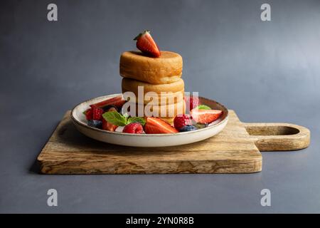 Ein Stapel flauschiger japanischer Pfannkuchen, serviert mit frischen Erdbeeren, Himbeeren, Heidelbeeren und Minzblättern auf einem rustikalen Holzbrett und Keramikplatte Stockfoto