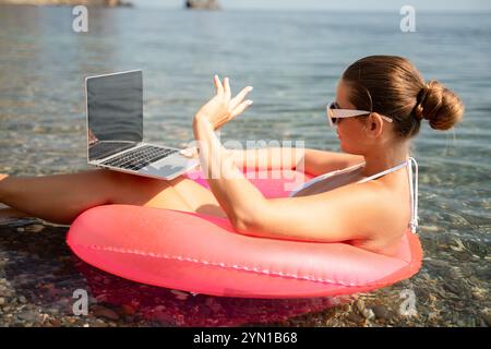 Eine Frau sitzt auf einem pinkfarbenen Schlauchboot im Wasser und benutzt einen Laptop. Das Konzept der Entspannung und Freizeit, während die Frau ihre Zeit am genießt Stockfoto