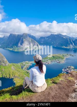 Eine Frau sitzt friedlich an einem grasbewachsenen Rand bei der Reinebringen Wanderung in Lofoten Norwegen und blickt über den atemberaubenden Fjord und die dramatischen Berge Norwegens. Stockfoto