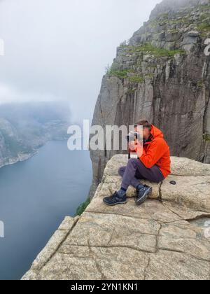 Auf einem felsigen Felsvorsprung von Preikestolen Norwegen konzentriert sich ein Fotograf in einer leuchtend orangefarbenen Jacke auf die atemberaubende Fjordlandschaft, die in Nebel gehüllt ist. Stockfoto