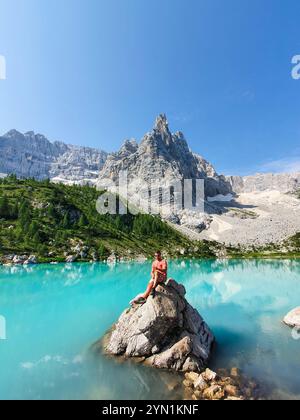 Eine Person entspannt sich auf einem Felsen an einem glitzernden türkisfarbenen See, umgeben von majestätischen Bergen und üppigem Grün, Lago Di Sorapis Dolomiten Italien Stockfoto