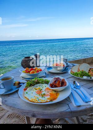 Genießen Sie ein köstliches Frühstück mit Eiern, Speck, frischen Salaten und Obst, das alle an einem Tisch mit Blick auf die ruhigen Meereswellen unter dem klaren blauen Himmel auf Aruba genossen wird Stockfoto