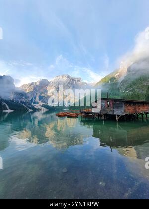 Am frühen Morgen umhüllt Nebel eine Holzhütte am Seeufer, die einen atemberaubenden Blick auf die Berge und das ruhige Wasser reflektiert und eine friedliche Atmosphäre in einer natürlichen Umgebung schafft. Lago Di Prags Dolomiten Italien Stockfoto