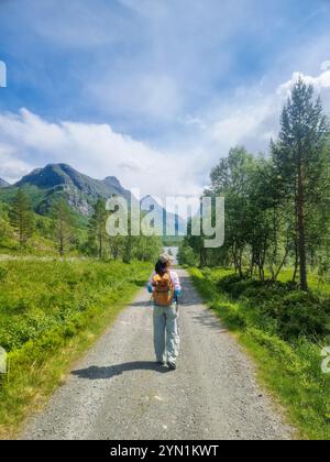 Ein Alleinreisender spaziert entlang eines ruhigen Schotterwegs in Innerdalen Norwegen, flankiert von lebhaftem Grün und majestätischen Bergen unter einem hellblauen Himmel, und genießt die Schönheit der Natur an einem perfekten Tag. Stockfoto