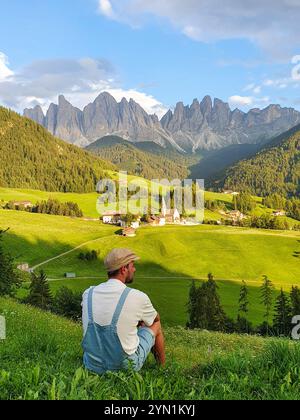 Ein Mann sitzt friedlich auf einem grasbewachsenen Hügel und genießt die atemberaubende Aussicht auf die majestätischen Dolomiten. Das lebhafte Grün und die ruhige Atmosphäre, Val Di Funes in Italien Stockfoto