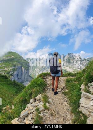 Ein Alleinreisender spaziert entlang eines felsigen Pfades, umgeben von atemberaubenden Bergen, unter einem hellblauen Himmel voller flauschiger Wolken. Die Zeit scheint still zu stehen in dieser ruhigen Landschaft der Schweiz Stockfoto