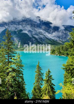 Ein atemberaubender türkisfarbener See reflektiert den blauen Himmel und flauschige Wolken, umgeben von hohen immergrünen Bäumen. Die imposanten Berge im Hintergrund. Oeschinensee Schweiz Stockfoto