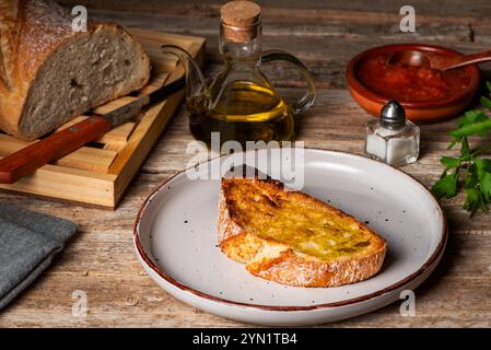 Toast mit Olivenöl neben einer Ölflasche, einer Schüssel mit gemahlenen Tomaten und einem Brotlaib, rustikaler Holzhintergrund. Stockfoto